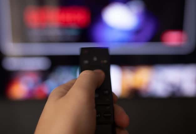 A hand holding a remote control pointing towards a TV screen displaying a cable guide with numerous channels and pricing information.