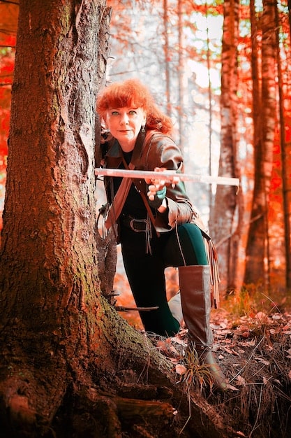 A cosplayer dressed as a warrior character, posed mid-jump in a forest setting. One leg is bent at the knee and raised high, while the other extends backward. The cosplayer is holding a sword overhead, with a determined expression on their face. Sunlight filters through the trees, creating dramatic lighting.