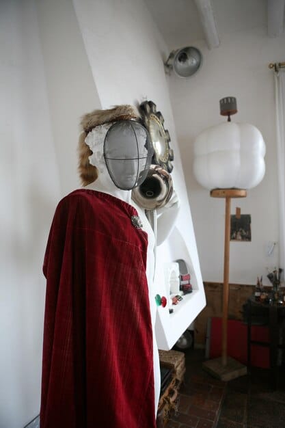 A cosplayer working on a large prop, wearing a respirator mask and using an airbrush to apply a base coat. The prop is partially painted, showcasing the smooth, even coverage achieved with the airbrush technique.