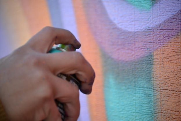 A hand carefully using an airbrush to apply a gradient of color to a light-colored cosplay wig. The wig is mounted on a styling head, and the background shows a well-lit crafting area with various tools and supplies.