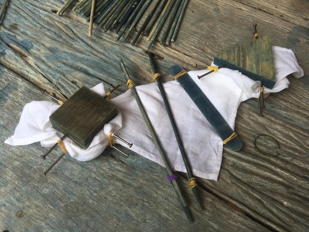 Close-up shot of a workbench showing various tools and materials used in cosplay weapon construction, including EVA foam sheets, cutting knives, heat guns, and bottles of acrylic paint. Shows attention to detail and safety measures.