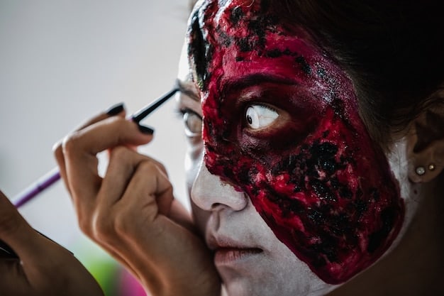 A crafter uses a small brush to apply fake blood to a latex appliance that mimics a gruesome wound on their hand. The setting includes various makeup tools and fake blood supplies.